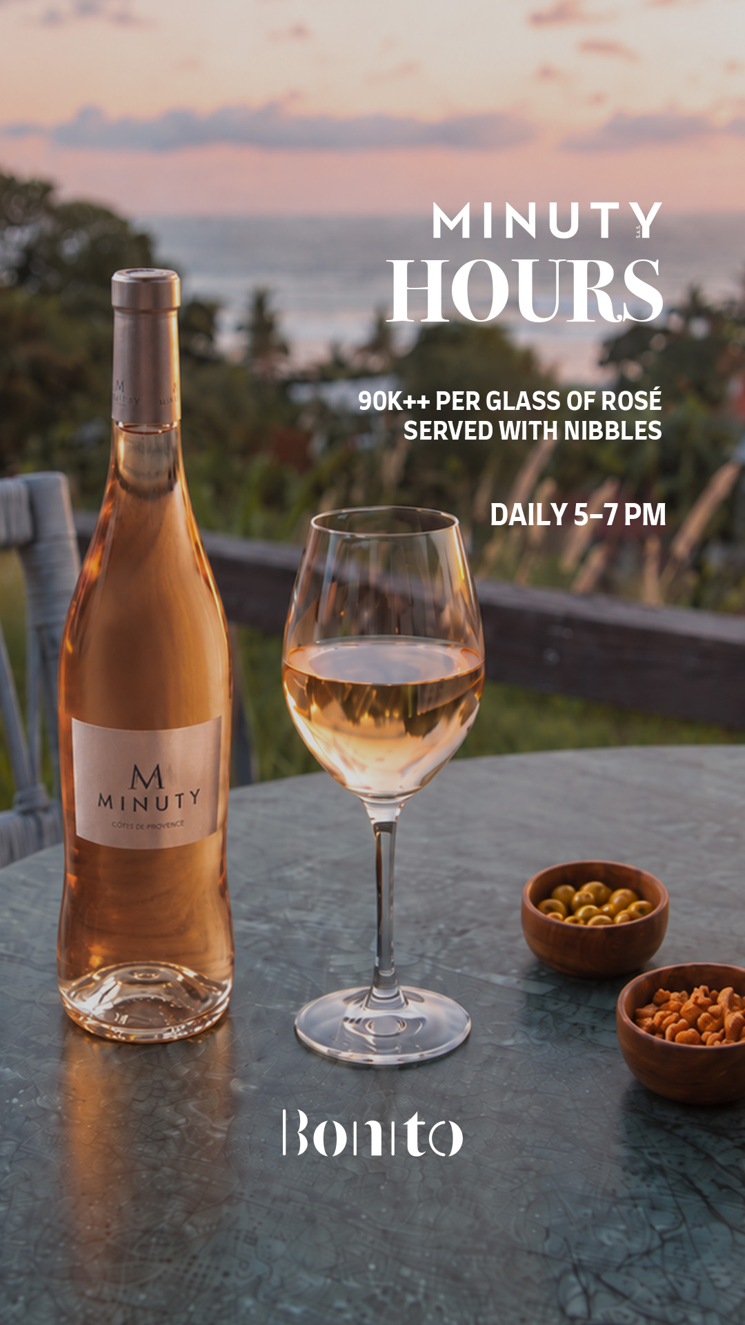 Bottle and glass of rosé wine on a table with bowls of olives and nuts, ocean view at dusk